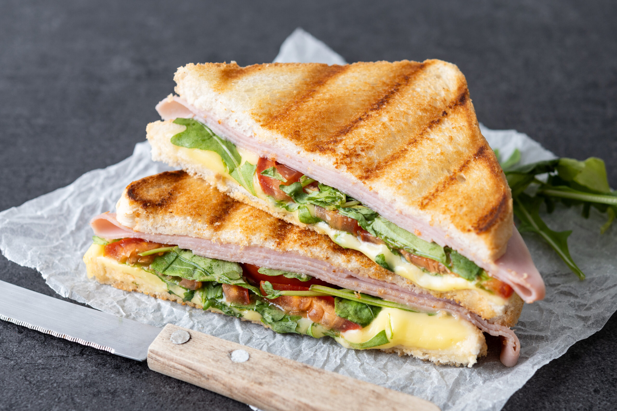 Panini sandwich with ham, cheese, tomato and arugula on black slate background